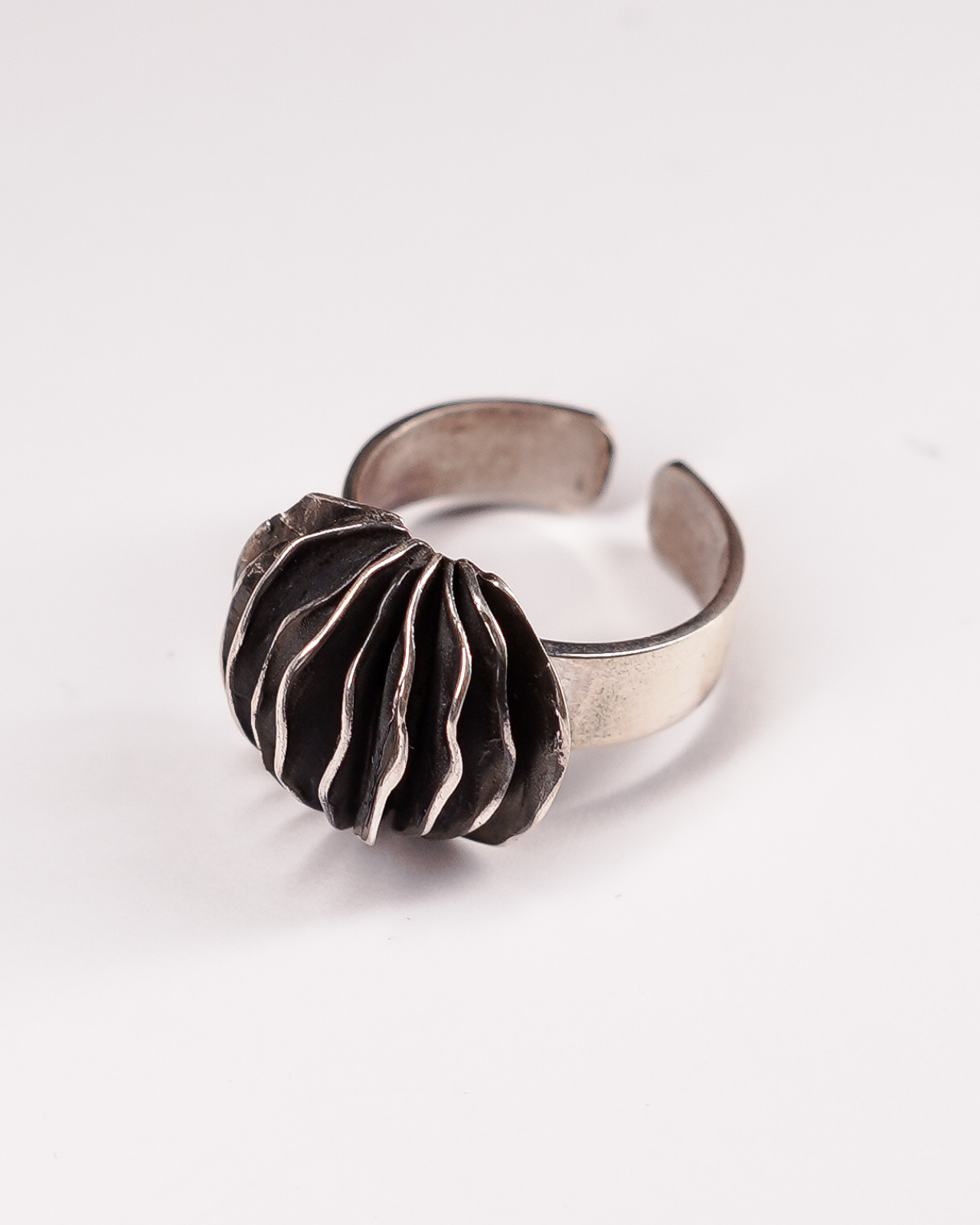 Anello regolabile in Argento 925 Éclat Gioielli Collezione Siwa, modello Guscio di Shali. Il design scultoreo presenta lamelle sottili e ondulate sovrapposte con finitura brunita negli incavi e profili lucidi, evocando una forma organica e stratificata. Gioiello artigianale su sfondo bianco."