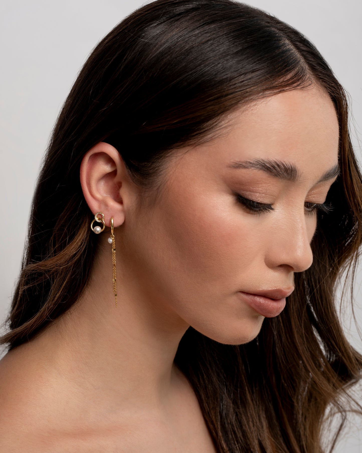 Gros plan d'une femme aux longs cheveux bruns portant une boucle d'oreille pendante dorée avec de fines chaînes et une petite perle, visible sur l'oreille droite." éclat gioielli