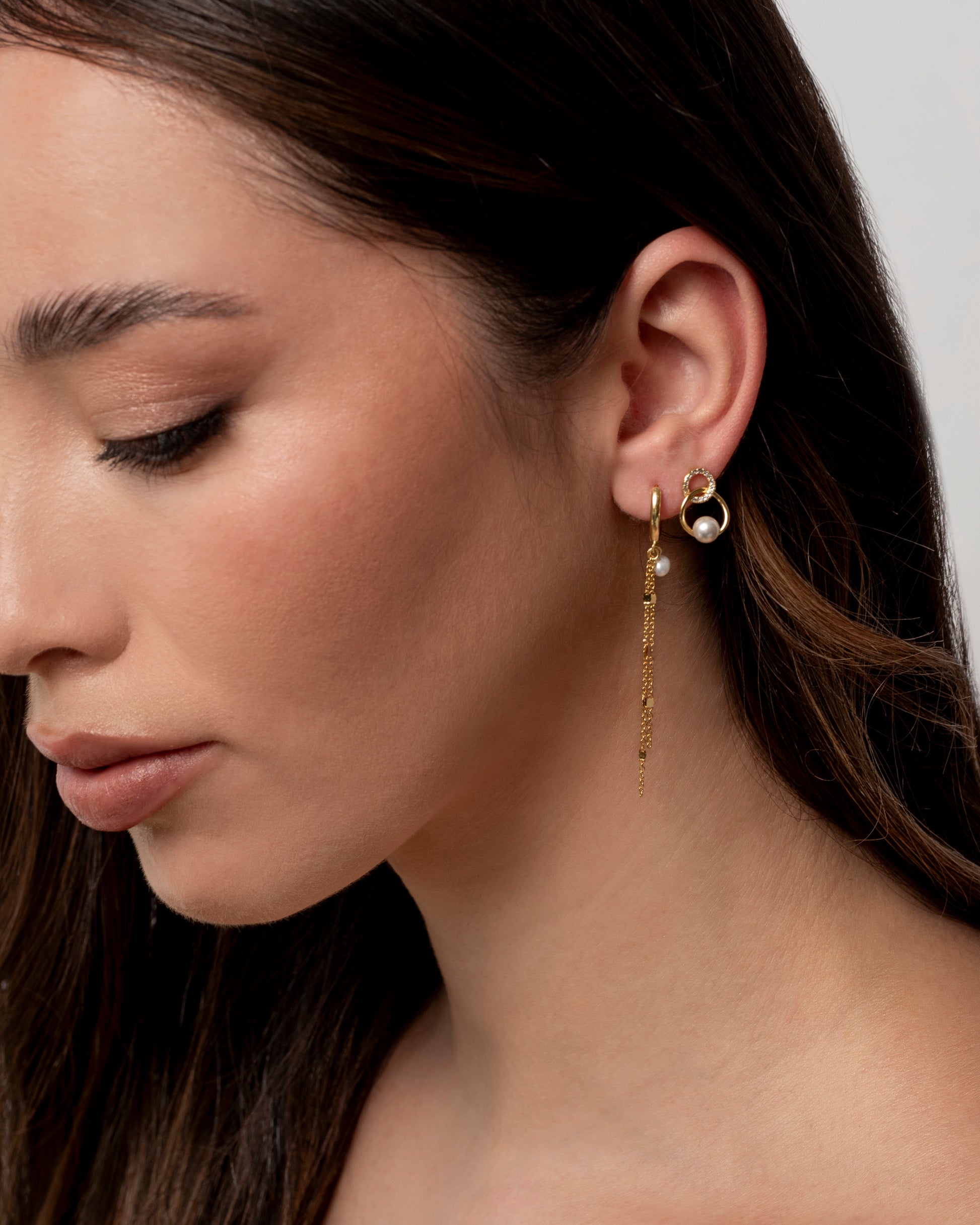 lose-up di una donna con lunghi capelli castani che indossa un orecchino pendente color oro con catenine sottili e una piccola perla, visibile sull'orecchio destro. eclat gioielli