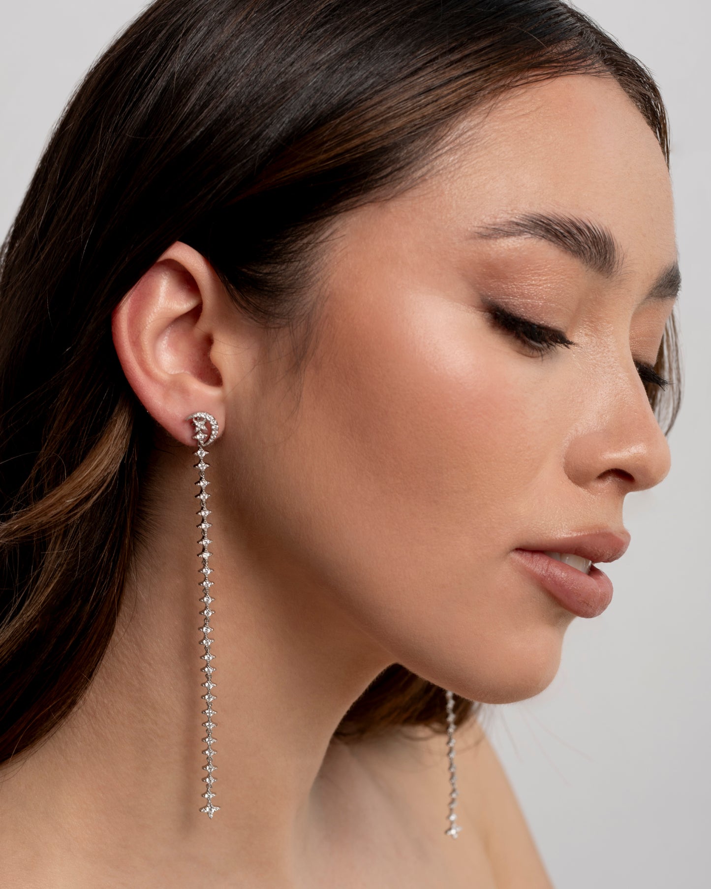 Close-up di una donna che indossa un orecchino pendente lungo e scintillante della collezione "Cascata di Luna" di Éclat Gioielli. L'orecchino è composto da un elemento superiore a mezzaluna e una cascata di piccole pietre preziose, che scende elegantemente. La donna ha capelli scuri e un trucco leggero, mettendo in risalto il gioiello.