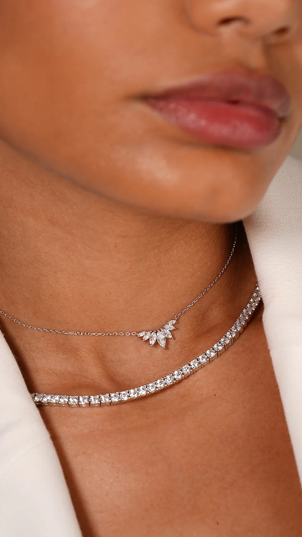Primo piano di donna che indossa due collane, una collana tennis 3mm in argento 925 con zirconi brillanti e una collana sottile con ciondolo a foglia, su giacca bianca. Gioielli Eclat.