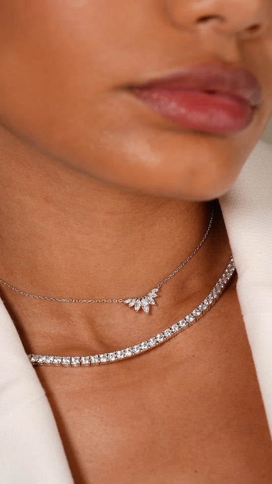 Primo piano di donna che indossa due collane, una collana tennis 3mm in argento 925 con zirconi brillanti e una collana sottile con ciondolo a foglia, su giacca bianca. Gioielli Eclat.