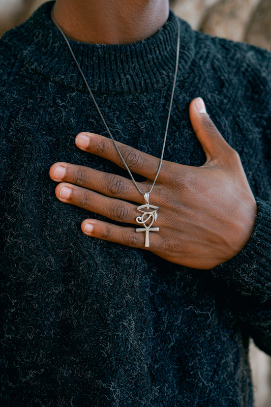 Ciondolo egizio in Argento 925 che unisce l'Ankh e l'Occhio di Horus, indossato su una catenina. Primo piano sul gioiello e sul maglione a trecce scuro.