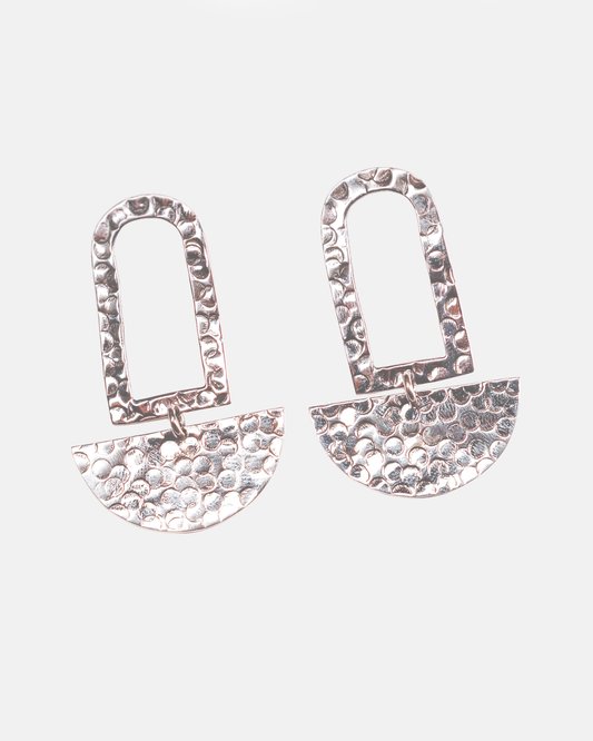 Coppia di orecchini pendenti in Argento 925 martellato a mano Éclat Gioielli Collezione Siwa. Il design presenta una forma a arco geometrico superiore e un elemento a semicerchio martellato inferiore che ricorda una luna calante, su uno sfondo grigio chiaro pulito. Gioielli artigianali in argento martellato."